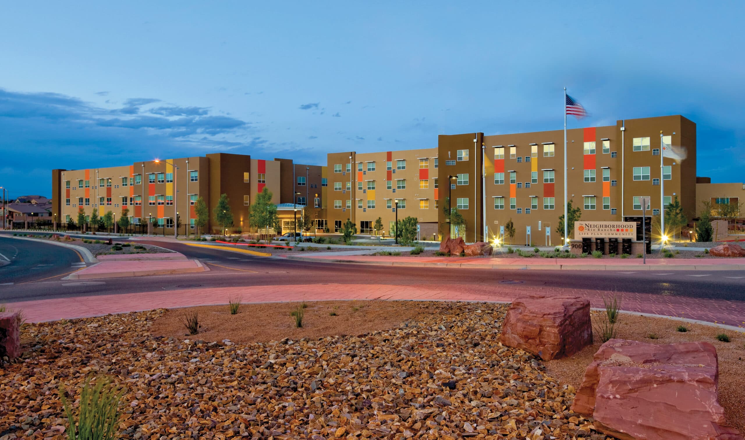 Wide exterior view of the Neighborhood Rio Rancho life-plan community building at twilight, with warm lights glowing through its windows and an American flag flying above the entrance.