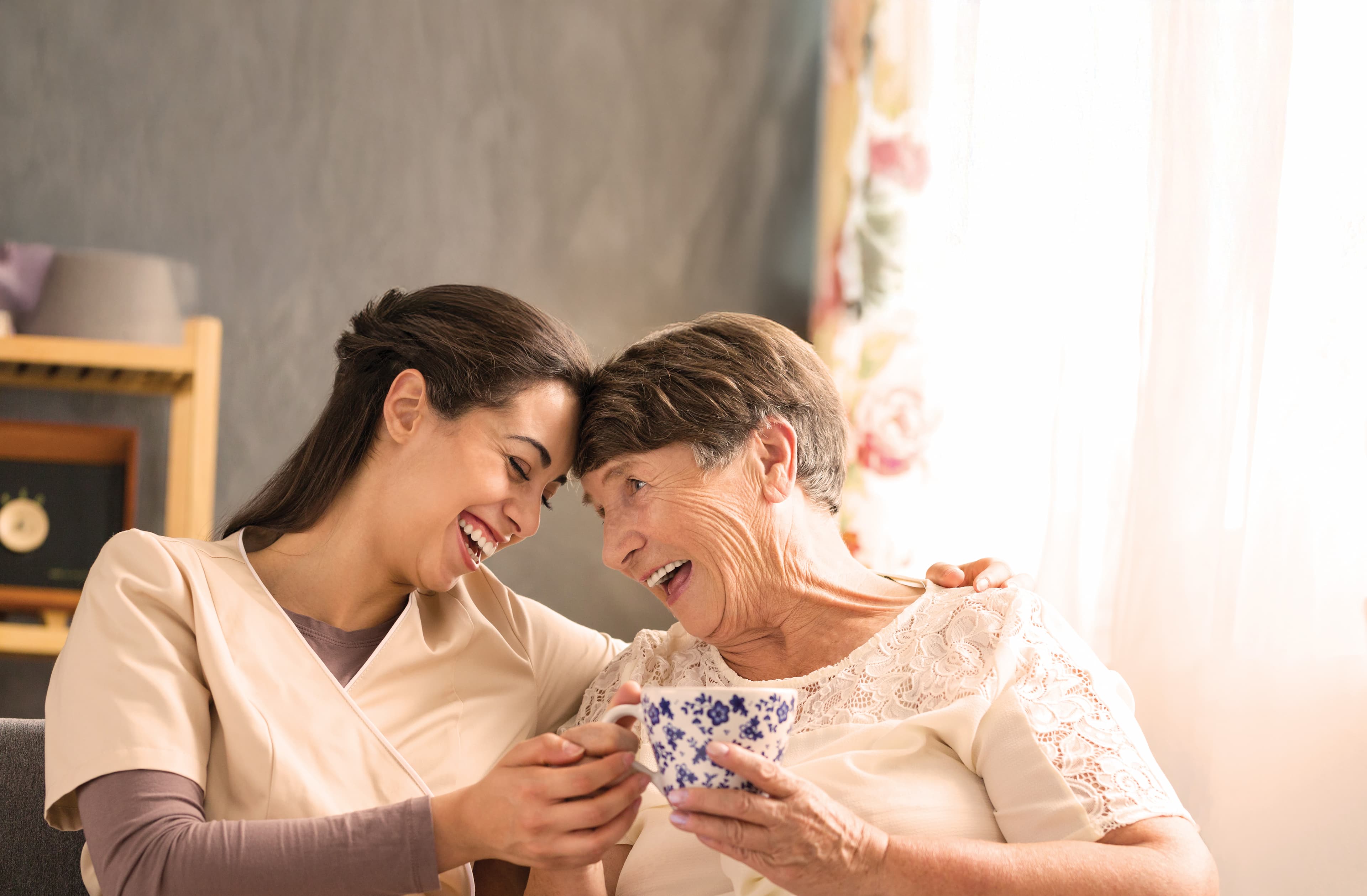 Caregiver and older woman share a laugh while sipping tea in warm window light.